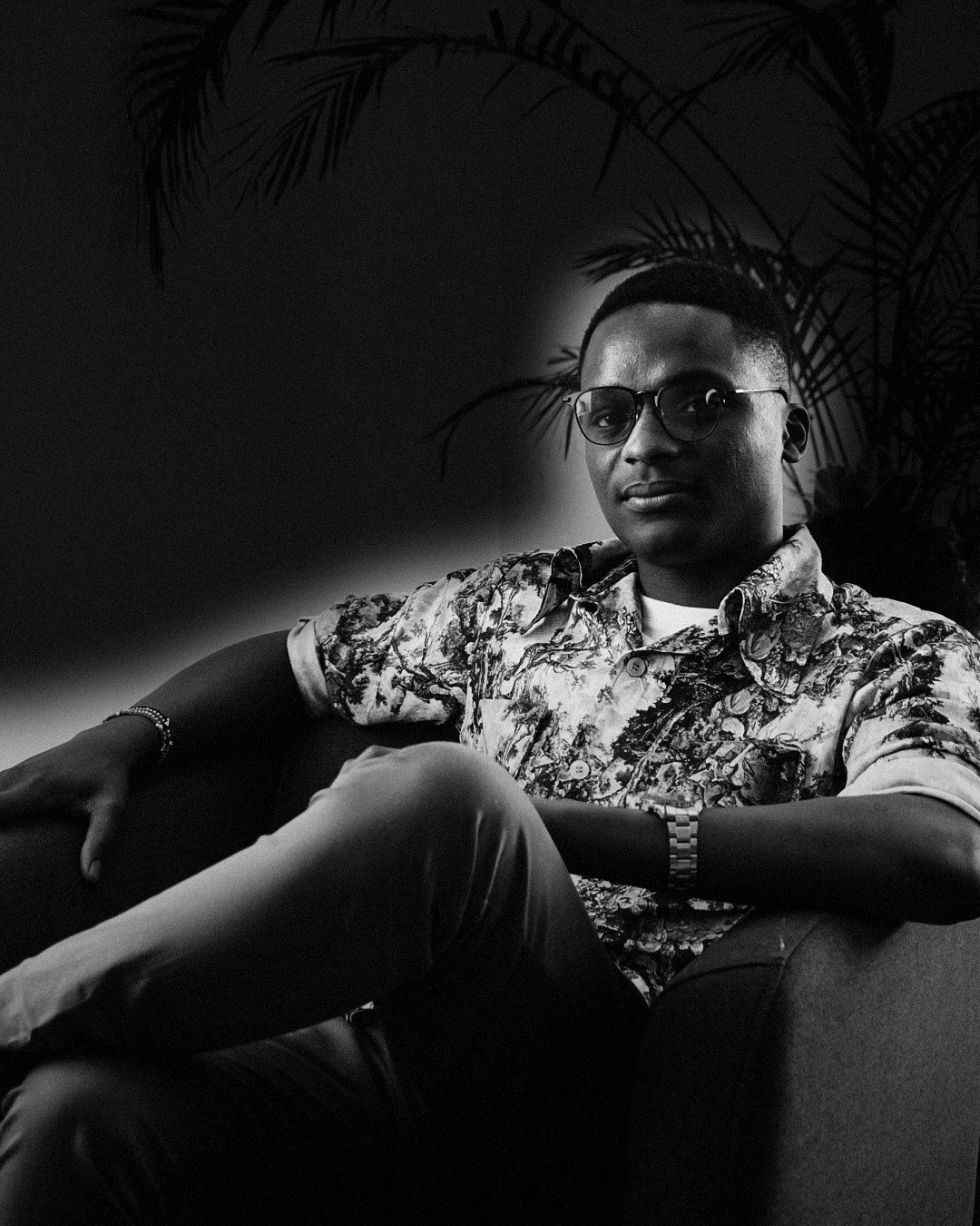 Black-and-white portrait of Chris Okoth seated in an armchair, in glasses and a patterned shirt